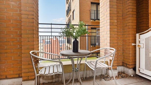 A small outdoor balcony with a round table and two chairs, a potted plant, and a view of a brick building in the background. - London Choice -South Kensington-Museums (London)