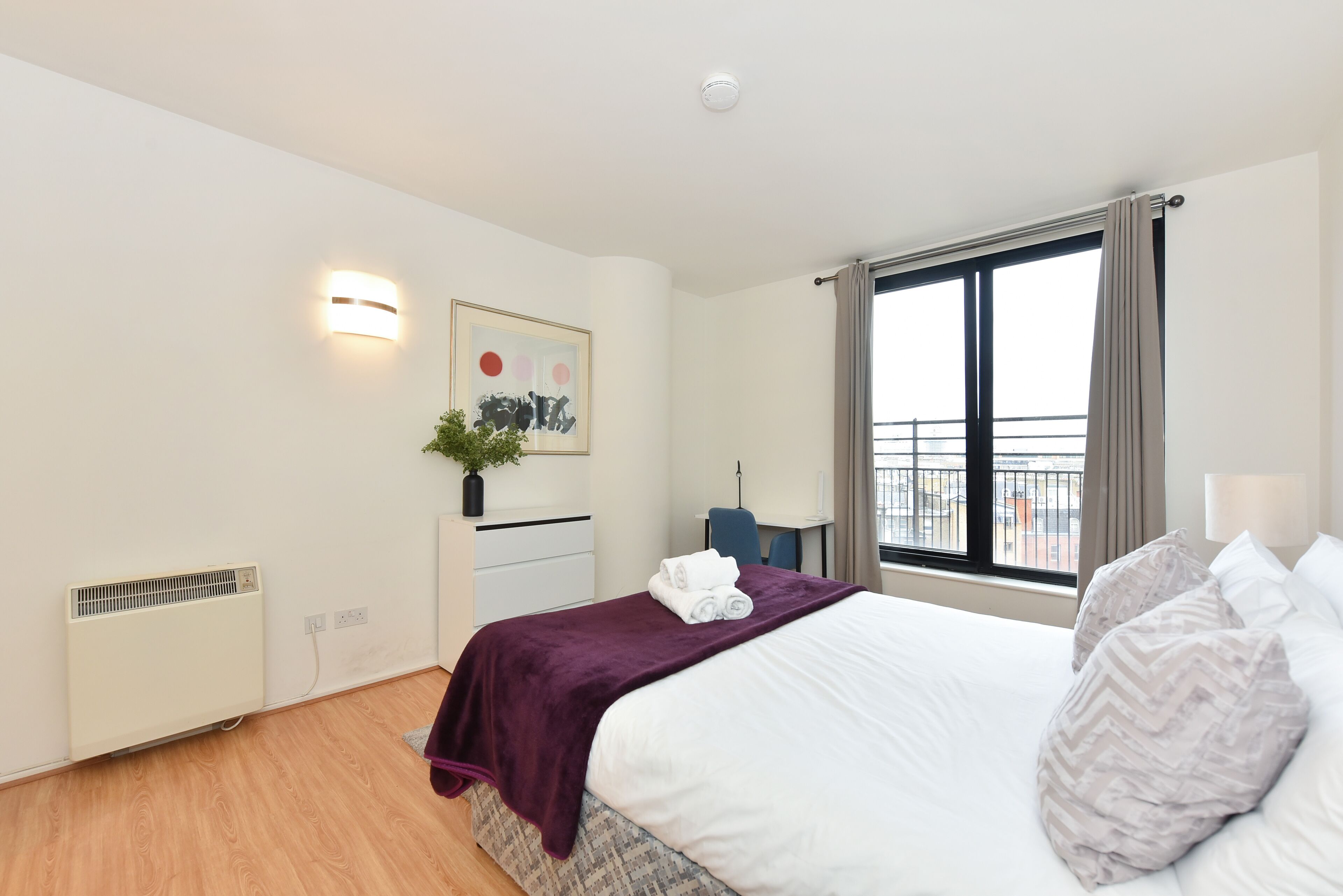 A modern hotel room with a neatly made bed, a radiator, a desk with a chair, and a large window with curtains.