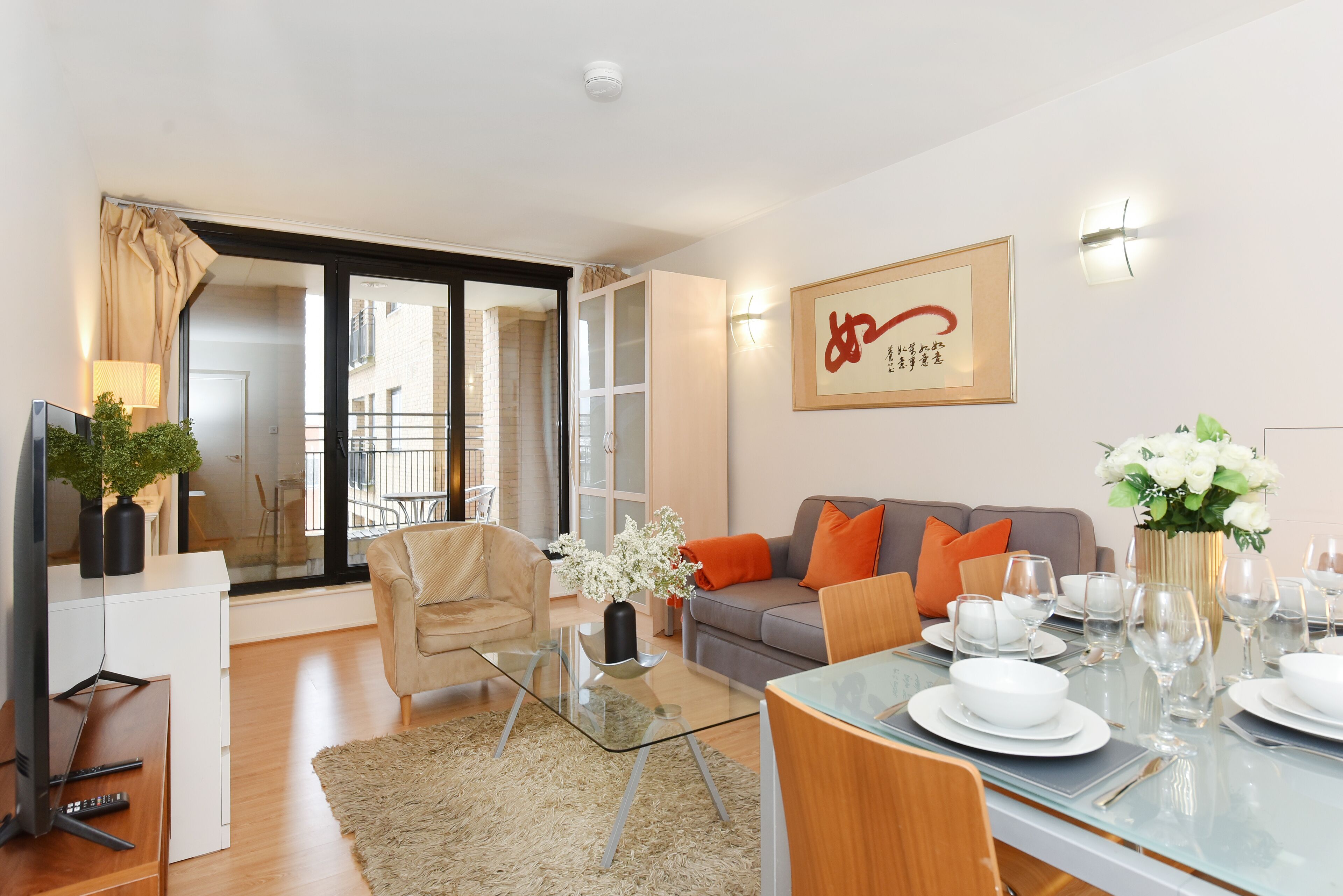 Een moderne woonkamer met een glazen salontafel, een bank met oranje kussens, een eettafel gedekt voor vier personen, en een kunstwerk in een lijst aan de muur.