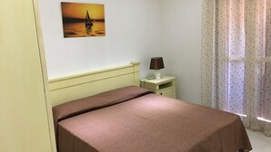 A bedroom with a bed, a nightstand with a lamp, a painting of a sunset over water, and a window with curtains. - TORREGRANDE BEACH ORISTANO (Torre Grande)