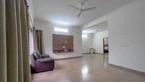 Un salón amplio con sofá, televisor montado, ventilador de techo y un baño visible al fondo.