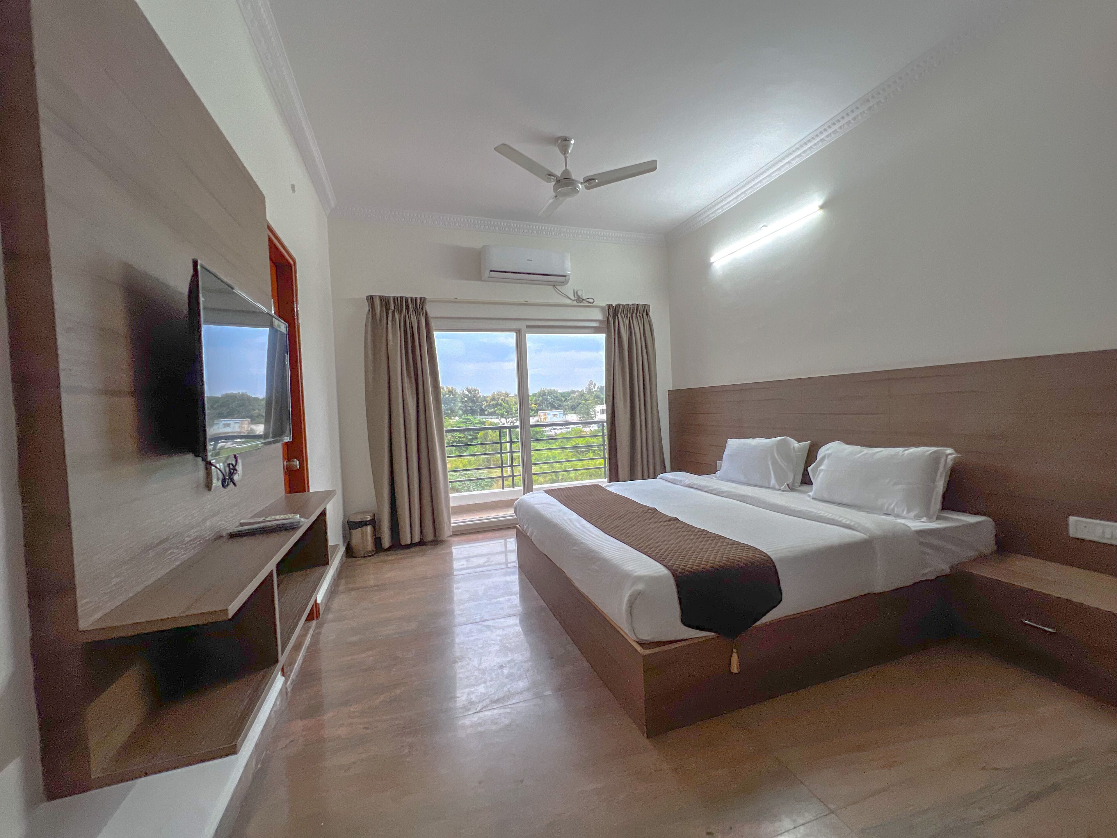 A hotel room with a large bed, a TV mounted on the wall, a ceiling fan, and a view of the outdoors through a large window.