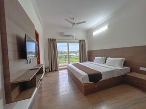 Habitación de hotel con una cama grande, un televisor montado en la pared, un ventilador de techo y vistas al exterior a través de un amplio ventanal.