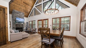 A spacious living area with a fireplace, wooden ceiling, large windows, and a dining table.