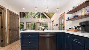 A modern kitchen with dark blue cabinets, a dishwasher, and a white countertop. There are hanging light fixtures and a large window providing natural light.