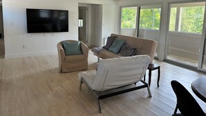 Amplia sala de estar con una televisión de pantalla plana montada en la pared, un sofá color beige con cojines, un sillón blanco y una mesa de comedor con sillas. Amplias ventanas ofrecen luz natural y vistas al exterior.