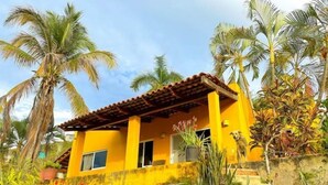 Une maison jaune avec un toit en tuiles, entourée de palmiers et d’un jardin où poussent différentes espèces de plantes.