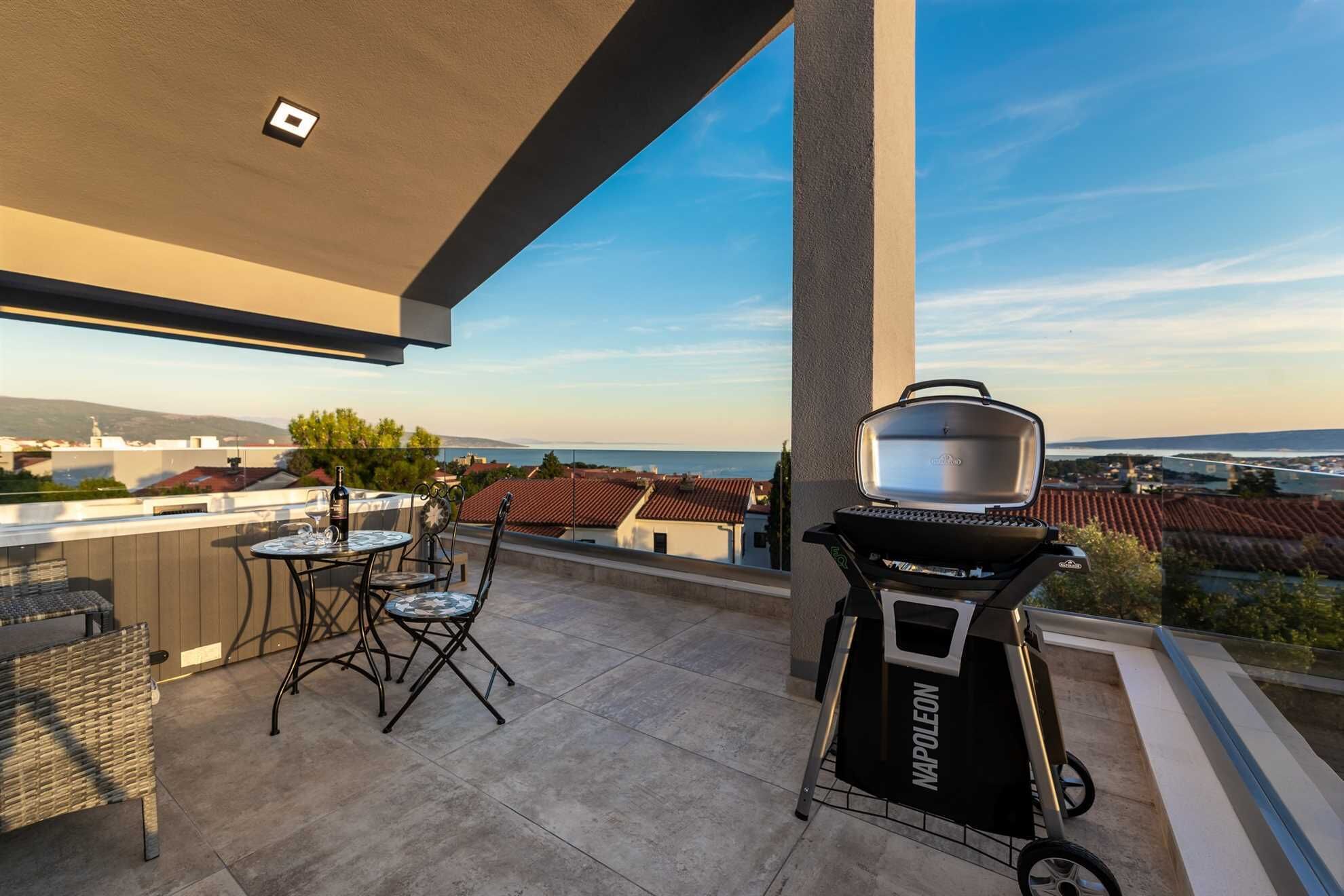 A rooftop terrace with a grill, a table with chairs, and a view of the city and sea.