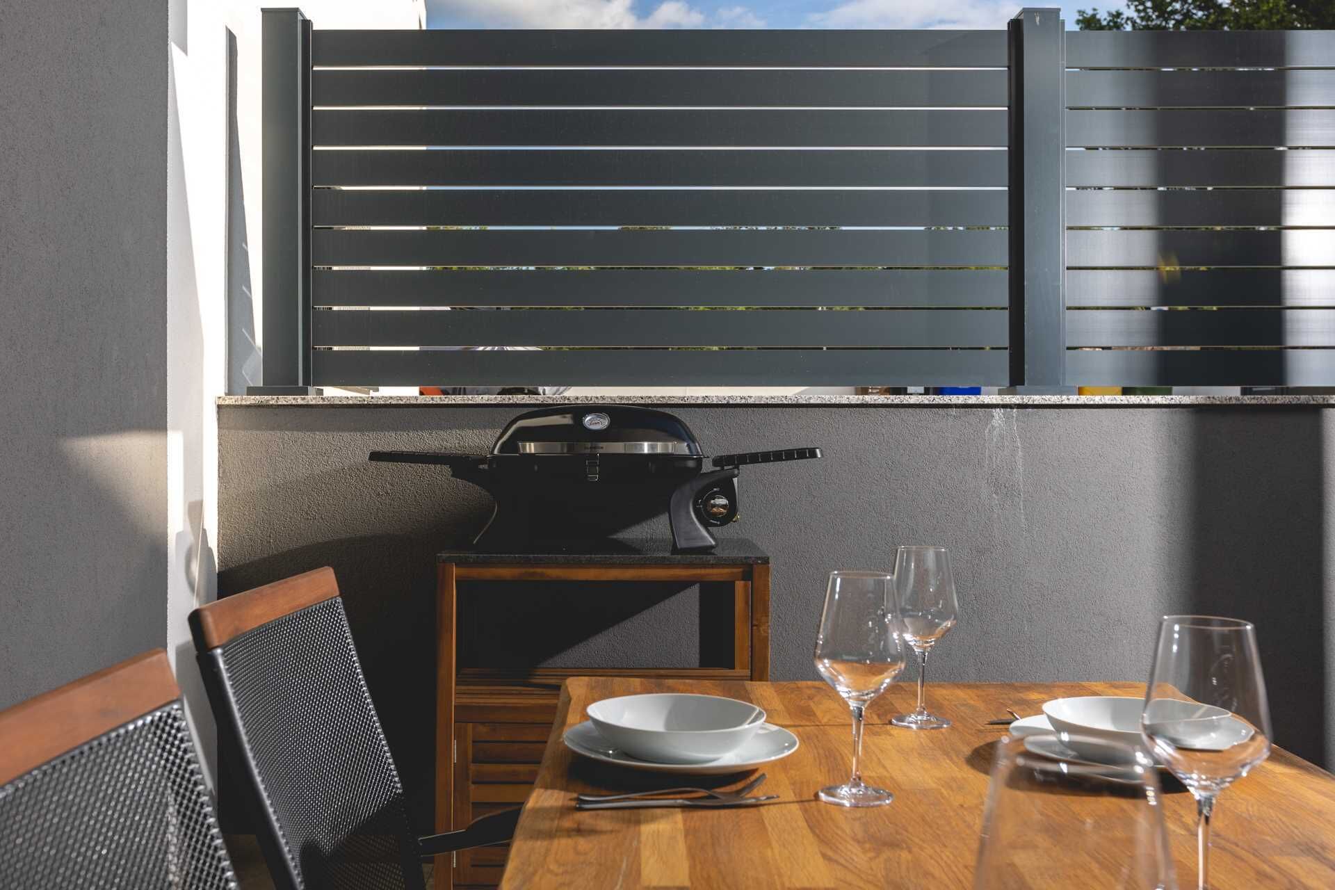 Ein Essbereich mit einem Holztisch für zwei Personen, einem Grill und Blick auf einen Zaun.