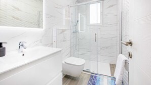 Ein modernes Badezimmer mit einer in Glas abgeschlossenen Dusche, weißen Marmorwänden und einem weißen Waschbecken inklusive Seifenspender.