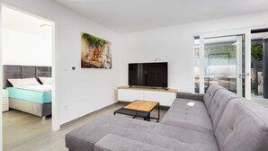 Ein modernes Wohnzimmer mit grauem Sofa, einem Flachbildfernseher, einem Gemälde und einem Essbereich mit Tisch und Stühlen.