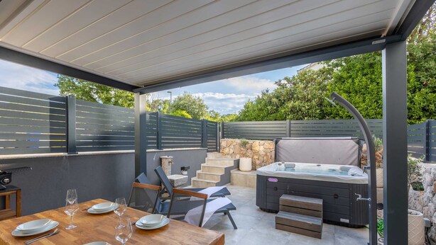 Ein überdachter Patio-Bereich mit Esstisch für vier Personen, Sitzgelegenheiten im Freien, einem Whirlpool und einer Wand aus Stein.