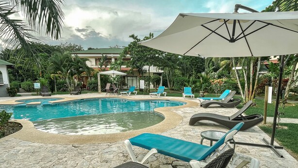 Zona de piscina con sillas de descanso y sombrillas, rodeada de vegetación y edificios.