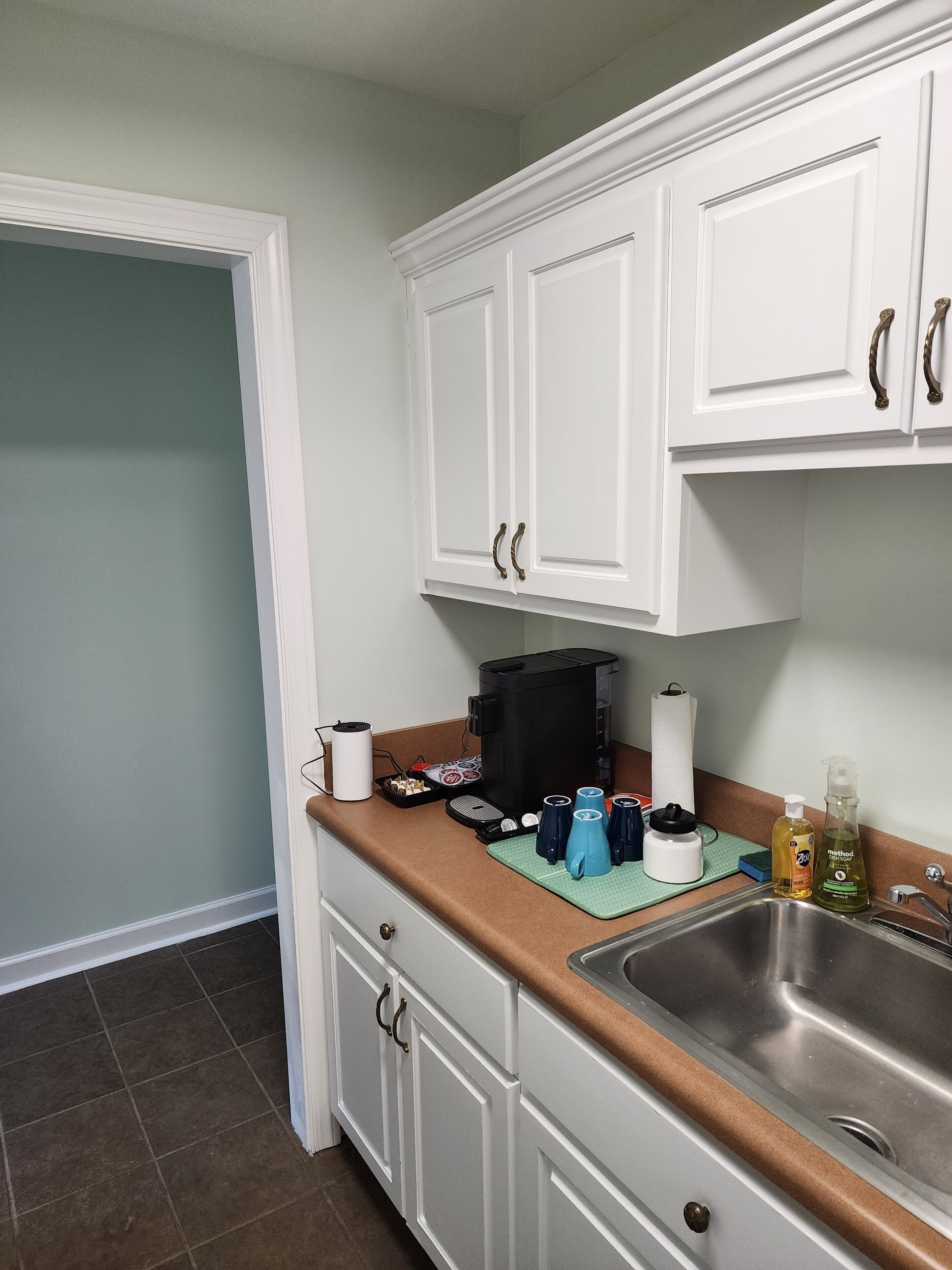 A kitchen with white cabinets, a stainless steel sink, and a countertop with various items including a coffee maker, a kettle, and a dish rack.