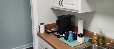 A kitchen with white cabinets, a stainless steel sink, and a countertop with various items including a coffee maker, a kettle, and a dish rack.