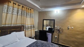 Une chambre d’hôtel équipée d’un lit, d’un bureau, d’une chaise, d’un miroir, d’un ventilateur sur pied et d’une lampe fixée au mur.