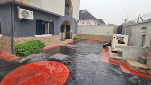 Un patio mojado con un motivo circular rojo, un edificio de fachada gris y una caja de servicios blanca.