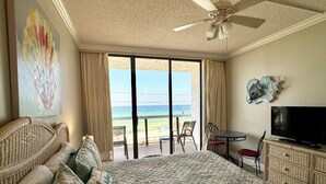 A hotel room with a large bed, a television, a balcony with a view of the beach, and a ceiling fan.