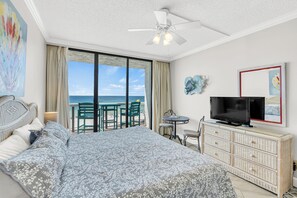 A bedroom with a large bed, a television, a dresser, and a balcony with a view of the sea.