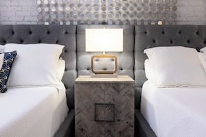 Une chambre d’hôtel avec deux lits, une tête de lit grise à motifs capitonnés, une table de chevet avec une lampe et un meuble aux motifs en chevrons.