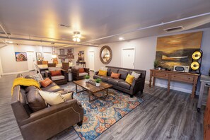 A spacious living room with a brown leather sofa, a wooden coffee table, and a colorful rug. There is a large painting on the wall and a wooden cabinet with a microwave. - Private Room #3 (Seneca)