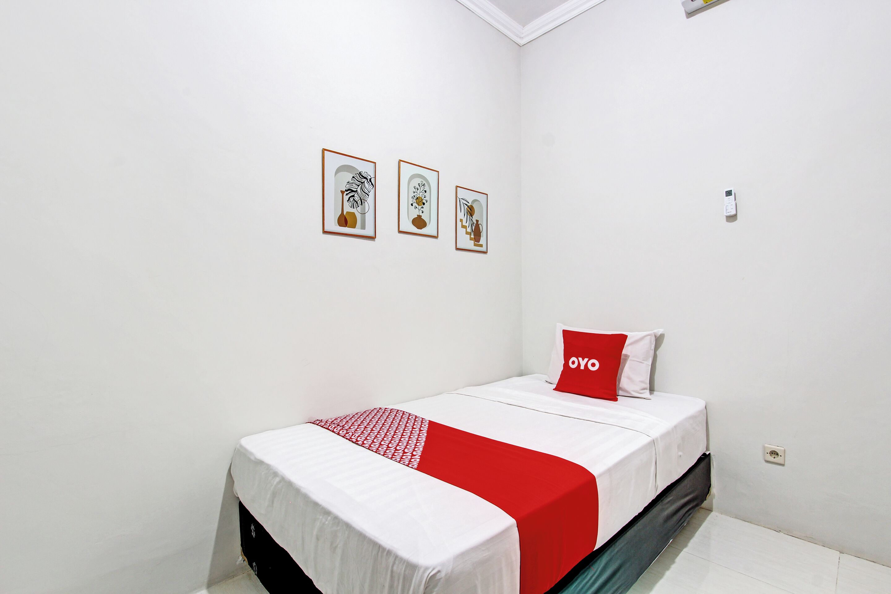 Une chambre bien rangée, avec un lit, un oreiller rouge et trois œuvres d’art encadrées accrochées au mur.