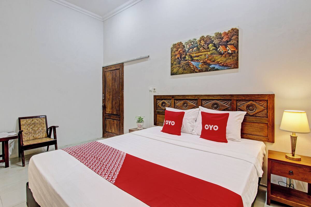 Ein ordentlich gemachtes Bett mit einer roten und weißen Bettdecke, zwei Kissen mit dem Schriftzug „OYO“, ein Holzkopfbrett, ein Landschaftsbild an der Wand, ein Stuhl, ein Nachttisch mit einer Lampe und eine Holztür.