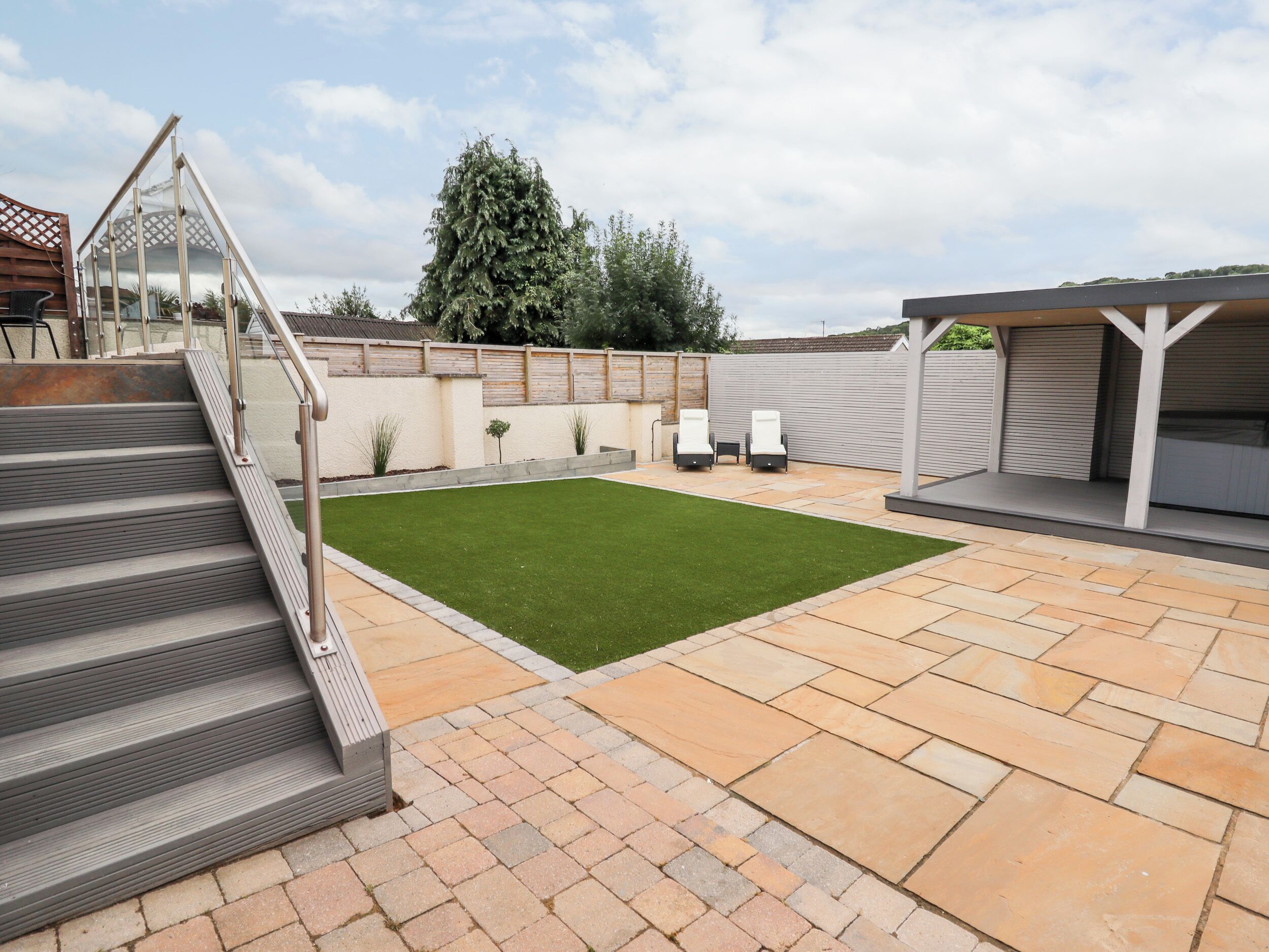 Un patio trasero con césped verde, una zona de patio con baldosas de piedra, un espacio cubierto para sentarse y una escalera que lleva a un nivel superior.