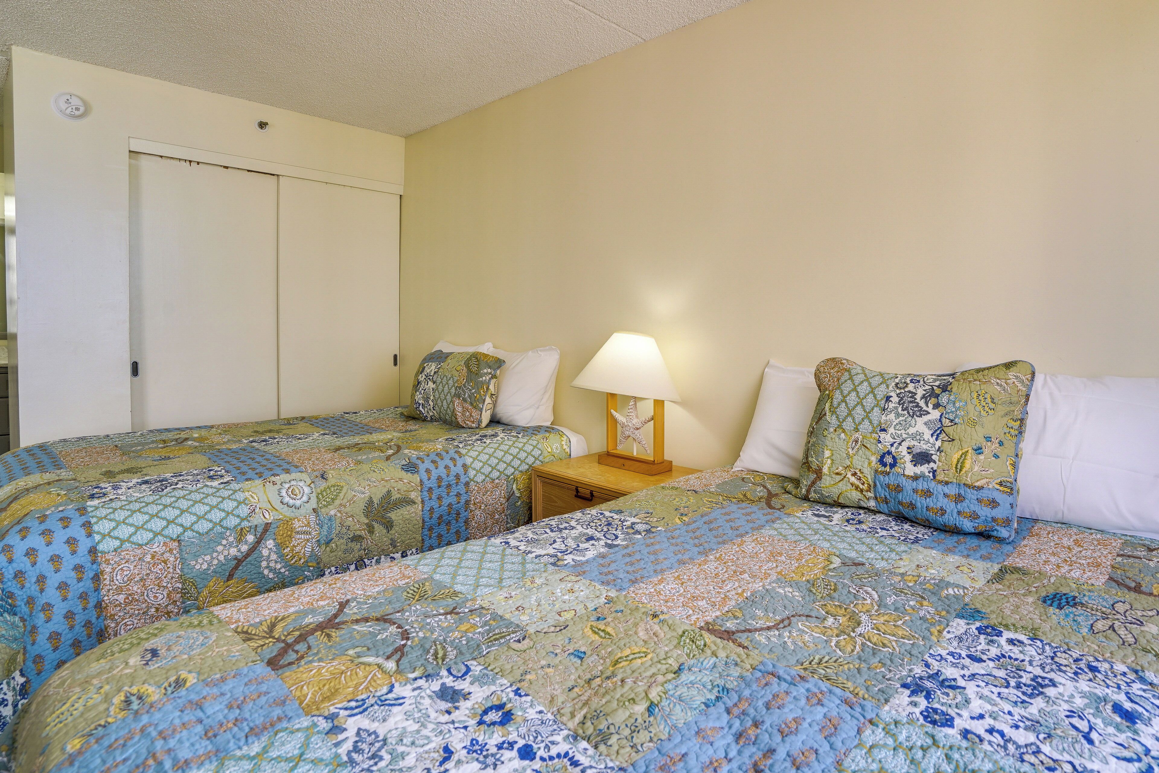 A room with two beds, each with floral-patterned bedding, a bedside table with a lamp, and a door.