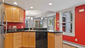A modern kitchen with wooden cabinets, a black dishwasher, and a large window with blinds.