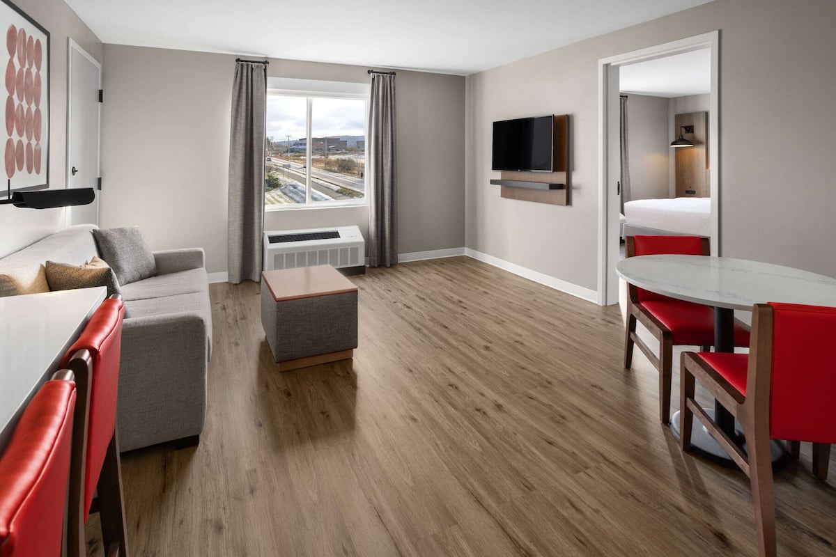 Une chambre d’hôtel moderne avec un canapé gris, un petit pouf, une table à manger avec des chaises rouges, un téléviseur à écran plat fixé au mur et une fenêtre donnant sur le paysage urbain.