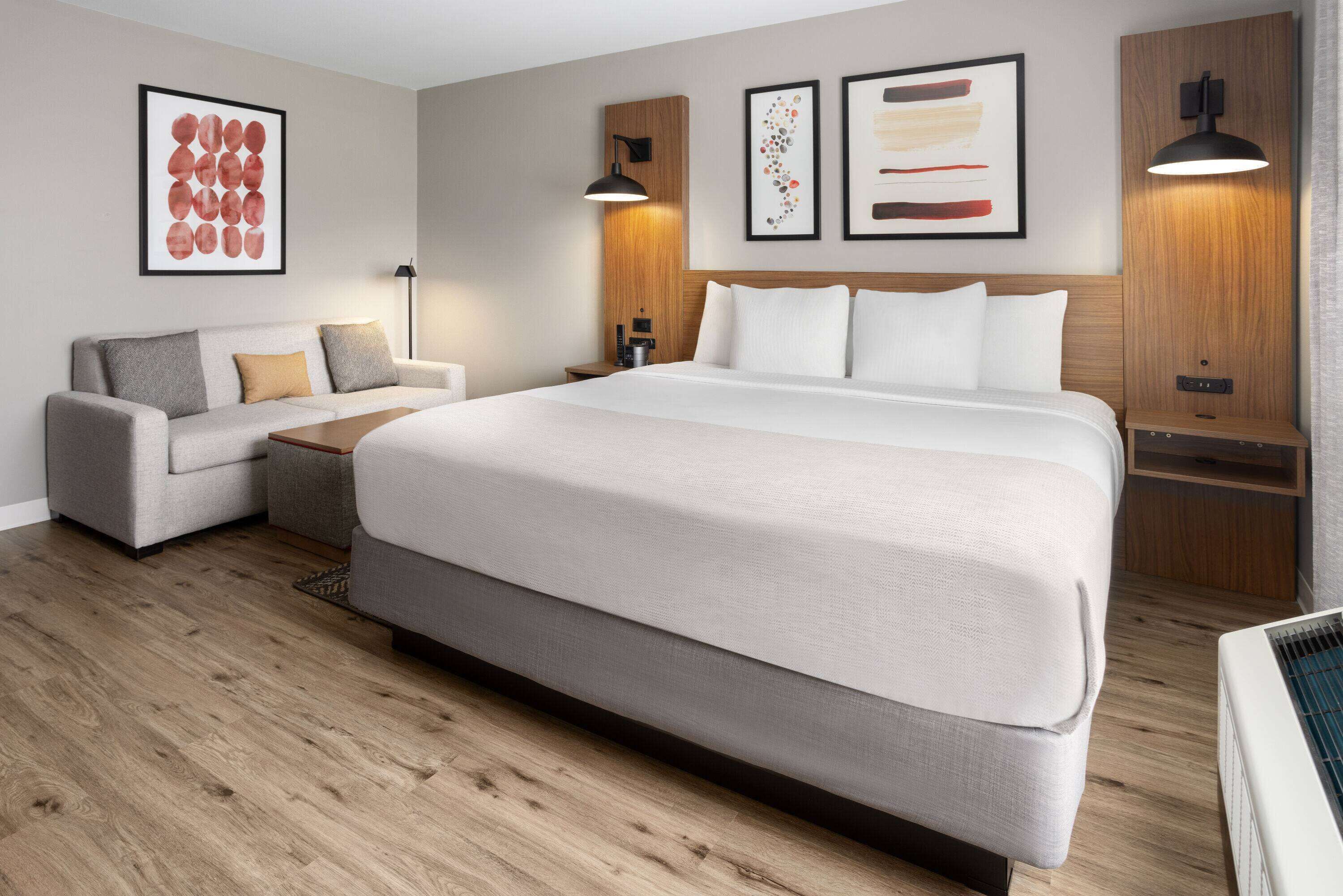 A modern hotel room with a large bed, a sofa, and a desk. There are wall-mounted lamps and framed artwork.