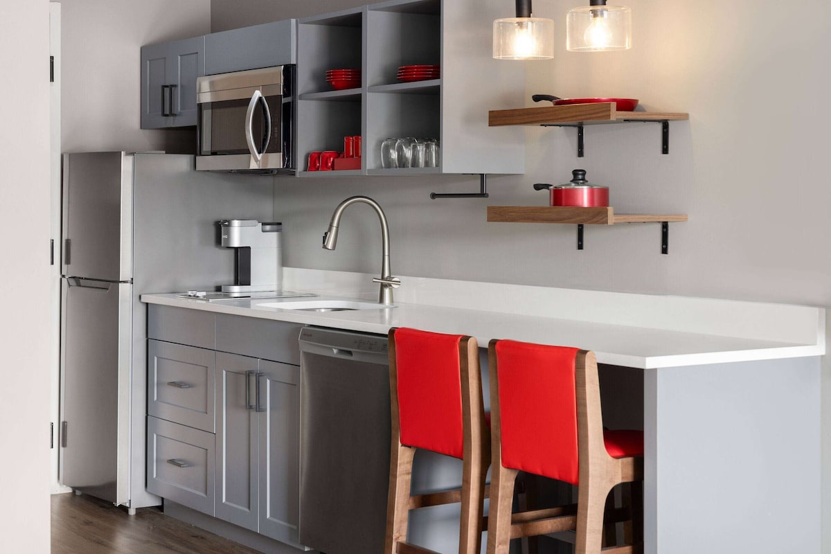 A modern kitchen with grey cabinets, a stainless steel refrigerator, a built-in oven, a sink, and a wooden chair with a red towel.