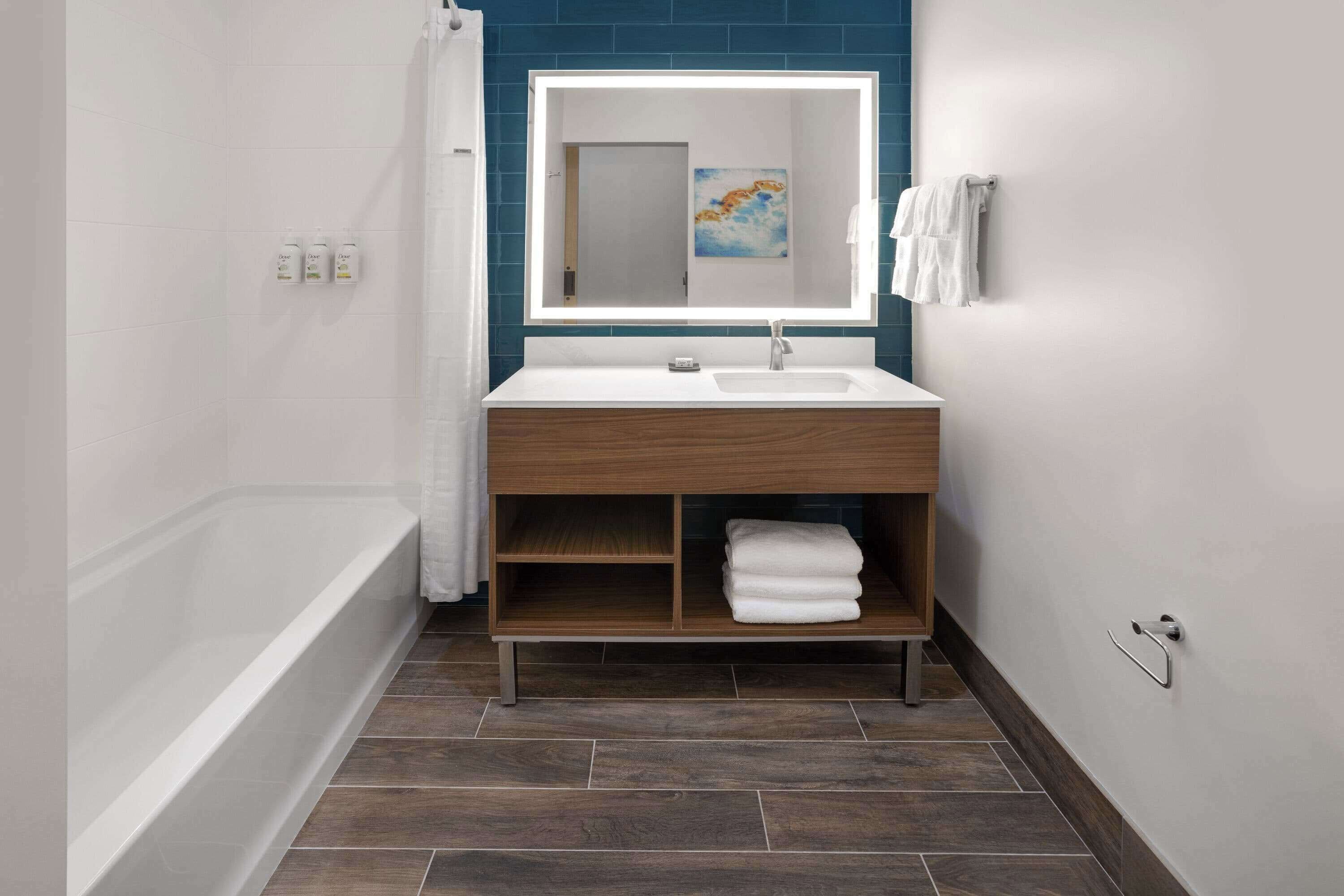 A modern bathroom with a white sink, a mirror with a light, a wooden vanity, a bathtub, and a towel rack.