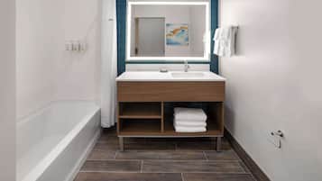 A modern bathroom with a white sink, a mirror with a light, a wooden vanity, a bathtub, and a towel rack.