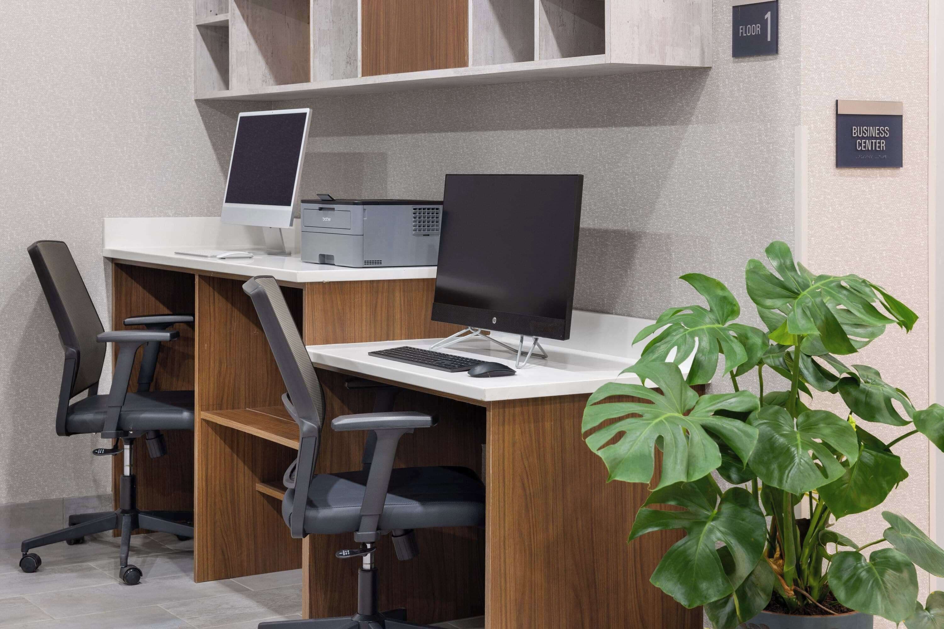 A modern office space with two desks, each with a computer and keyboard, and a printer. There are two office chairs, a potted plant, and signs indicating 'Floor 1' and 'Business Center'.