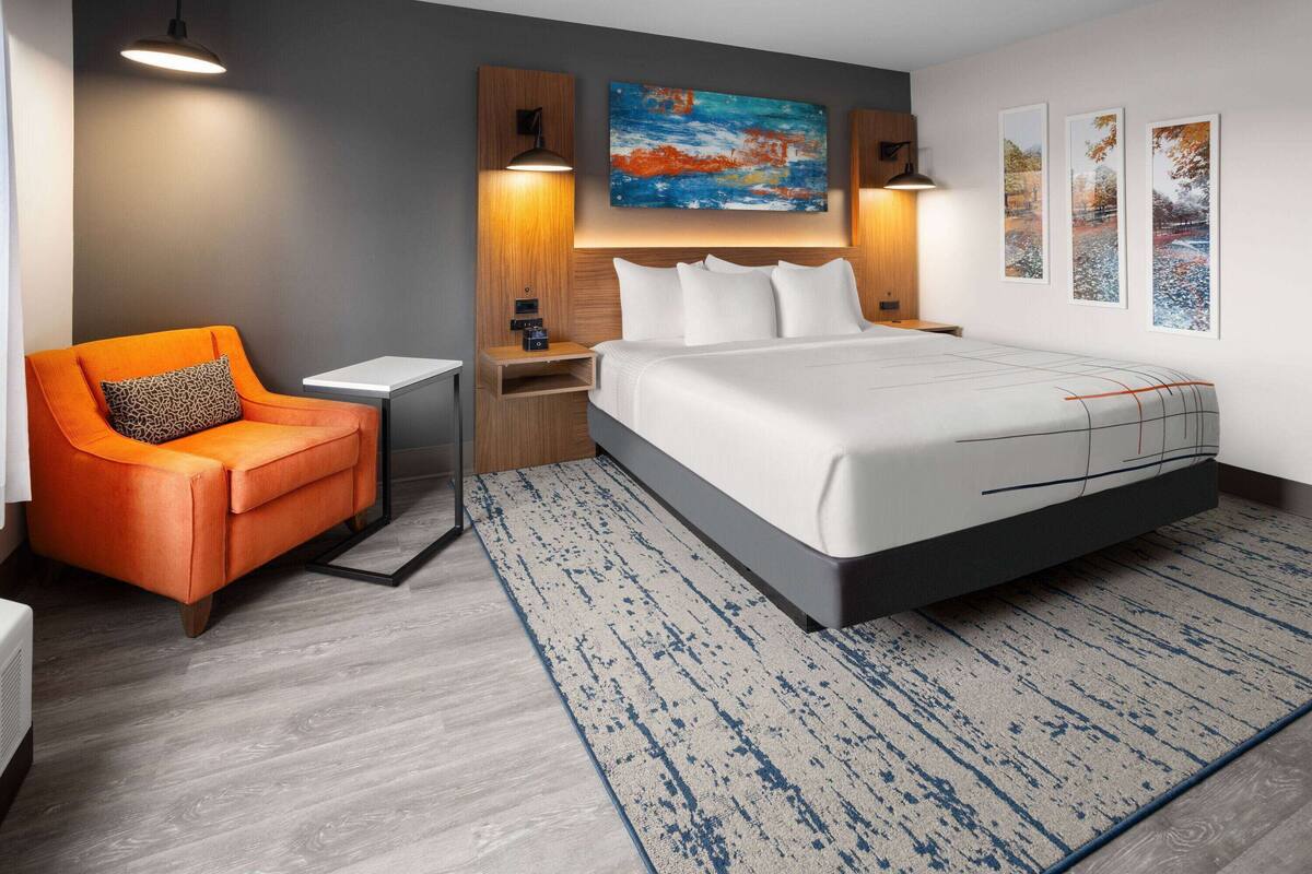 A modern hotel room with a large bed, a seating area with a chair, a nightstand, and a wall-mounted lamp.