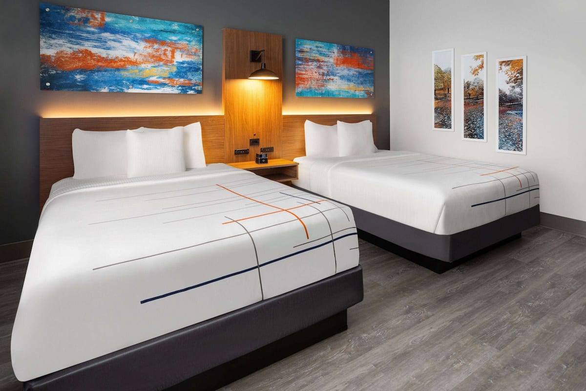 A hotel room with two beds, a wooden headboard, a nightstand, and three abstract paintings on the wall.