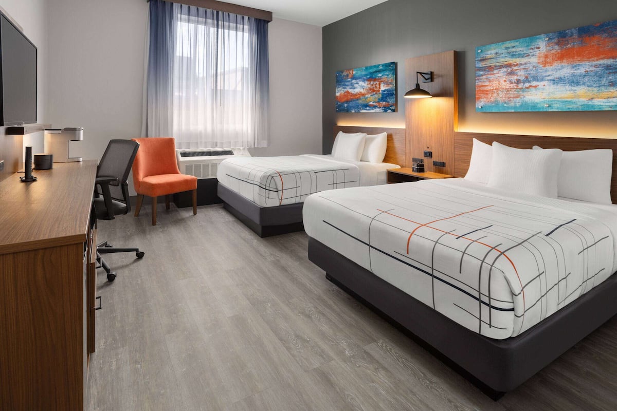 A hotel room with two beds, a desk, a chair, a TV, and artwork on the walls.