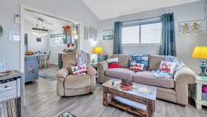 Een gezellige woonkamer met een bank, fauteuil en salontafel, een eetgedeelte met stoelen en een tafel, en een keuken met een koelkast en aanrecht.