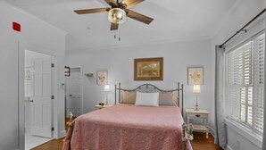 A bedroom with a wooden floor, a bed with a pink bedspread, two bedside tables with lamps, a ceiling fan, and a window with blinds.