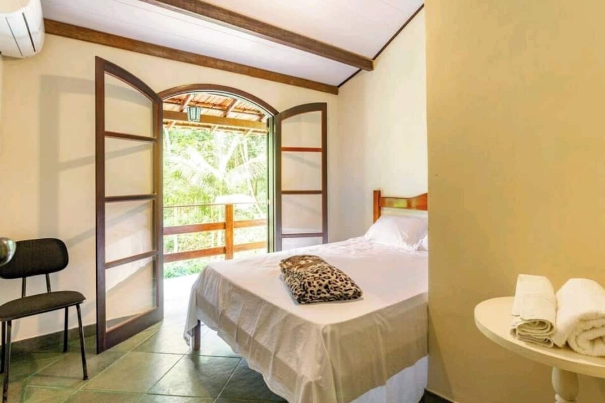 Quarto com cama, uma cadeira, uma mesinha com toalhas e vista para o verde através de uma porta aberta.
