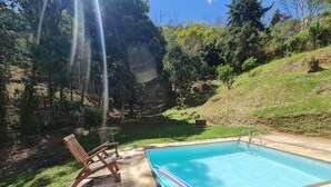 A swimming pool with a clear blue water, surrounded by a stone deck. There are two wooden chairs and a small table near the pool. - Sítio Borges  (Petrópolis)