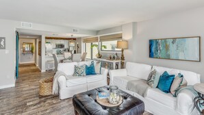 A modern living room with white sofas, a black ottoman, and a kitchen in the background. - Your Family Friendly Vacation Awaits! Olympic Size Pool! Short Walk to Beach (Pensacola Beach)