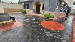 Un espacio exterior húmedo con suelo de baldosas, un diseño circular rojo y un edificio con balcón.