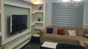 A modern living room with a flat-screen TV mounted on the wall, a beige sofa with cushions, a coffee table, and a chandelier. - Enjoy this unique, lavishly furnished 1-bed apartment with "Open Kitchen Design" (Lagos)