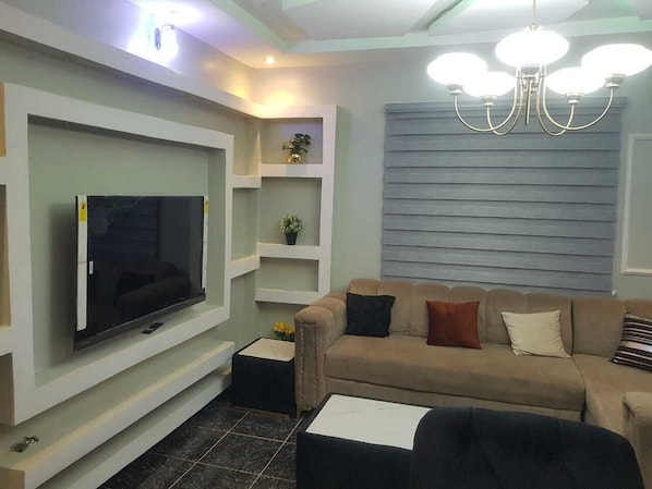 Un salon moderne avec un téléviseur à écran plat fixé au mur, un canapé beige agrémenté de coussins, une table basse et un lustre.