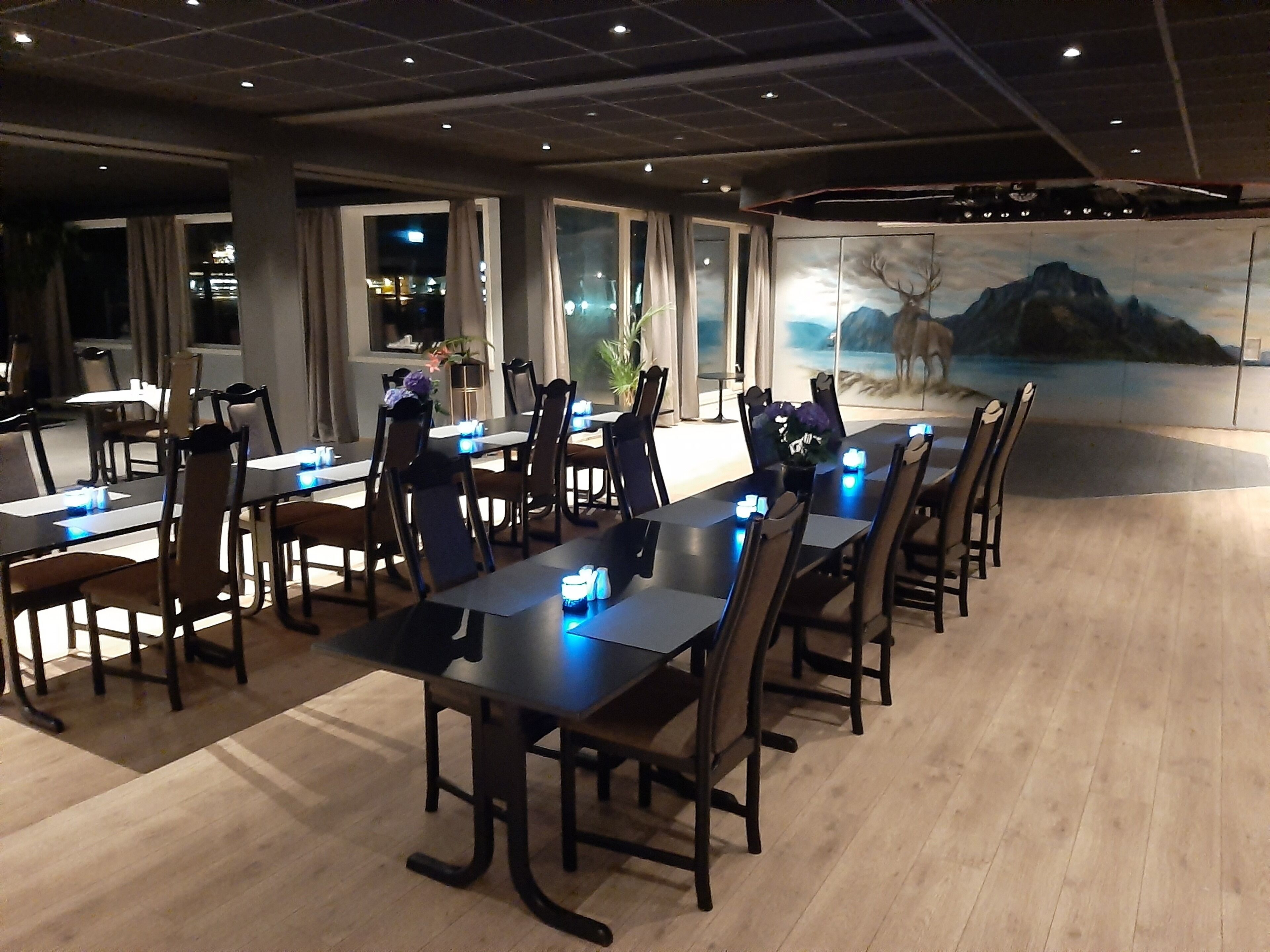 A conference room with long tables and chairs, a mural of a mountain scene, and a view of the city at night through large windows.