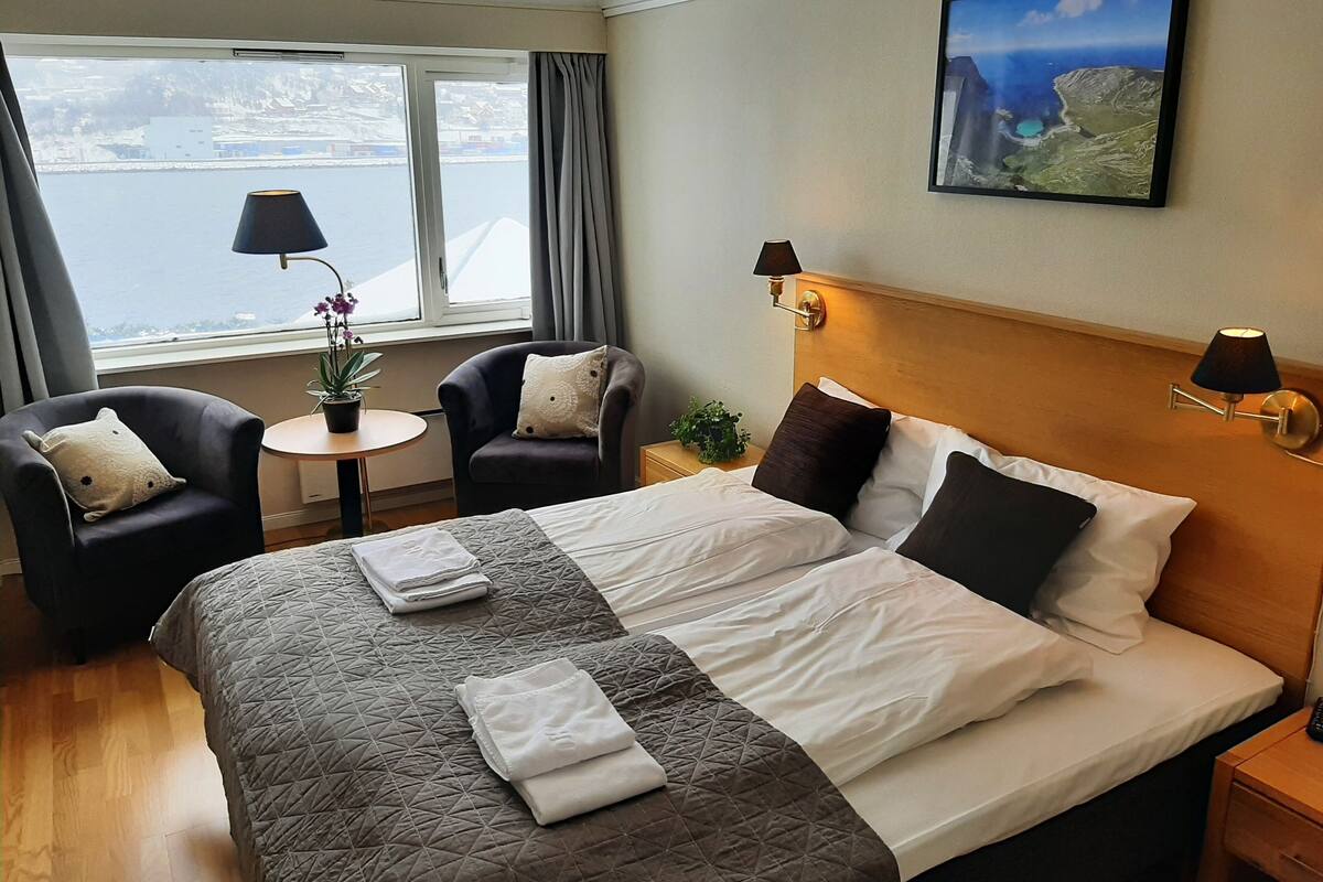 A hotel room with a large bed, two armchairs, a small table with a lamp, and a window with a view of the sea.