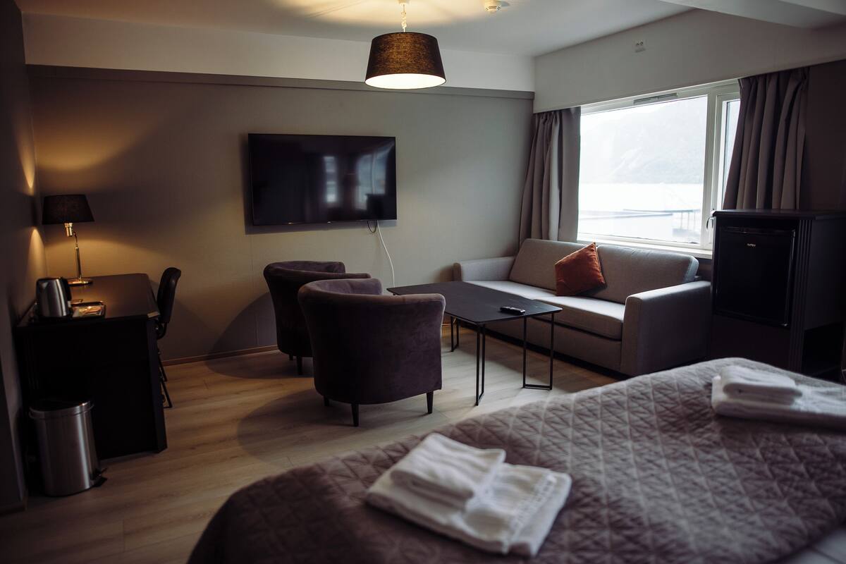 A modern hotel room with a bed, a sofa, a desk, a TV, and a window with curtains.
